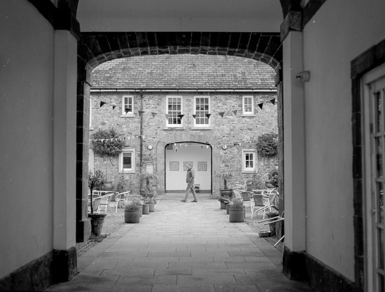 A black and white photo looking through an archway towards a stone building. Across the front of the building are dining chairs, with a man walking centre frame.