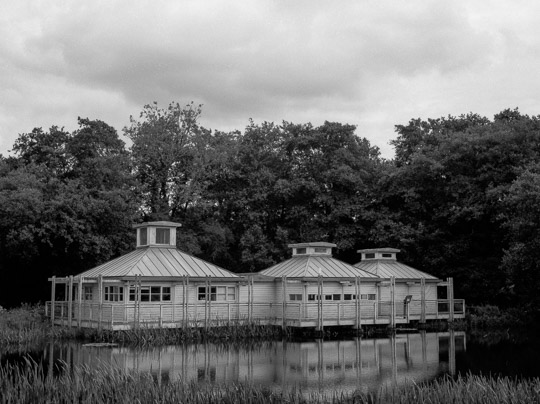 A black and white film photo of a building on stilts over a lake, with the building reflected in the mostly-still water below it.